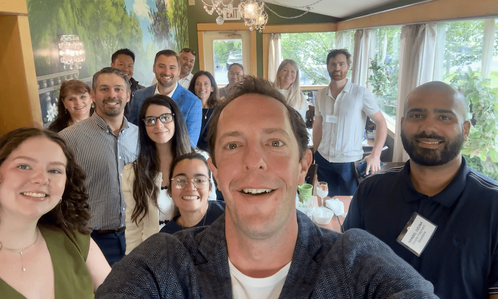 : A group selfie at Diana’s Café & Catering showing smiling panelists and attendees of the Executive Edge Food & Beverage event with Fitzpatrick. About a dozen people stand close together in a warmly lit room with large windows and a mural of trees in the background.