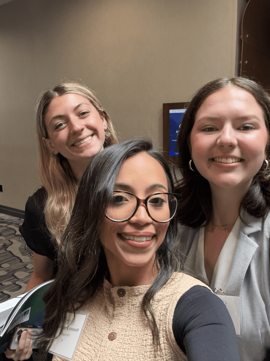 Modesty, Emily, and Peyton smiling for a selfie at a professional event, wearing name badges.