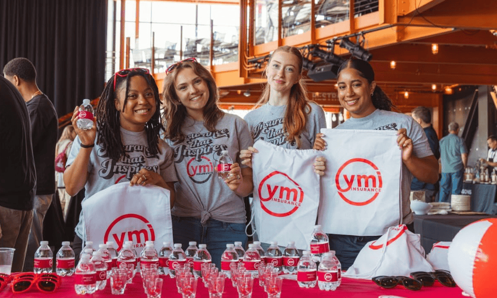 Four YMI team members pose behind the YMI giveaway table at the Ginvitational event at SteelStacks, showcasing branded drawstring bags, water bottles, sunglasses, shot glasses, and beach balls while welcoming attendees.